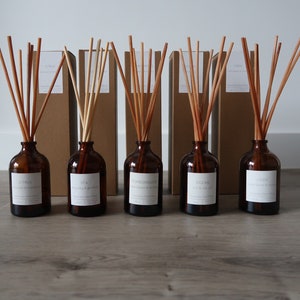 May include: Five brown glass bottles with brown wooden sticks in them. The bottles are labelled with different scents: Citrus, Spa, Pomegranate, Ocean, and Linen. The bottles are sitting on a wooden surface.