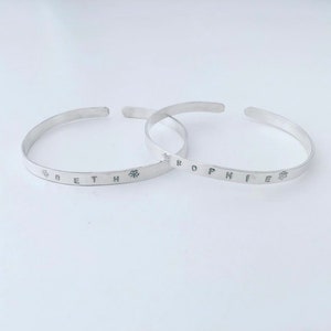 May include: Two silver bracelets with the names "Beth" and "Sophie" engraved on them. Each name is flanked by a small flower design.