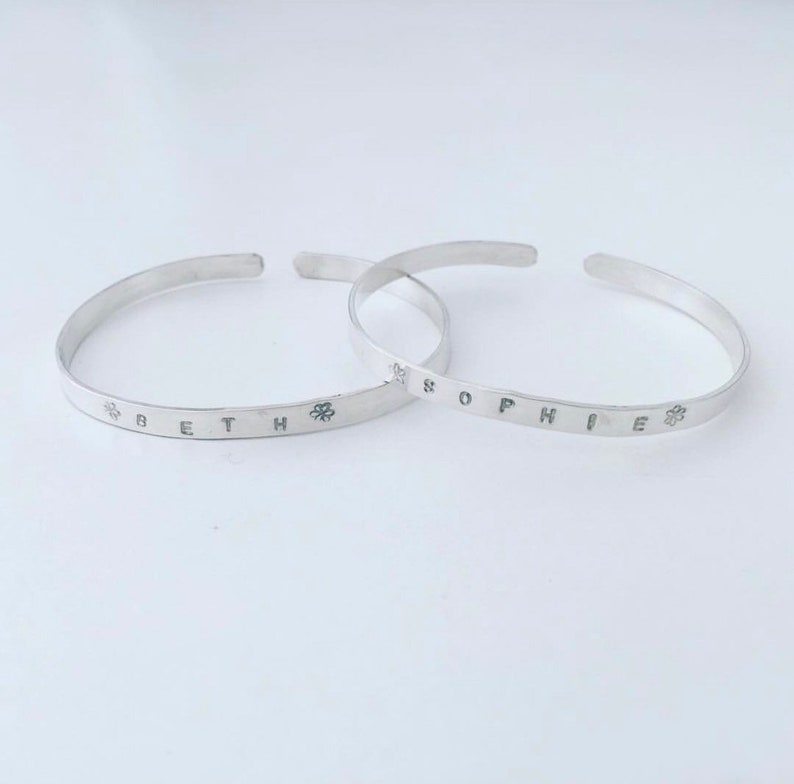 May include: Two silver bracelets with the names "Beth" and "Sophie" engraved on them. Each name is flanked by a small flower design.