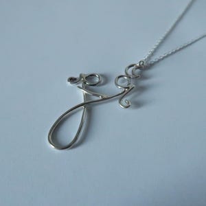 May include: A silver-colored necklace with a cursive "Joy" pendant. The pendant is crafted from wire and hangs from a delicate chain. The necklace is displayed on a light blue surface.