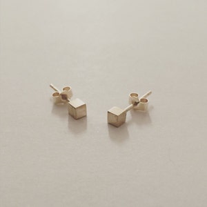 Könnte beinhalten: Ein Paar goldene Würfel-Ohrstecker. Die Ohrringe sind schlicht und minimalistisch, mit einem kleinen, quadratischen Würfel.