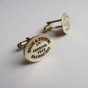 May include: A pair of gold-colored cufflinks with an oval face. The face of each cufflink has the text "WALLIS & STEEVENS LTD ENGINEERS 7943 BASINGSTOKE" in black. The cufflinks are on a white background.