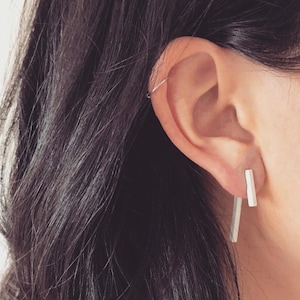 May include: A pair of silver earrings with a minimalist design. The earrings are long and thin, with a rectangular shape. They are worn in the earlobes.