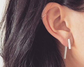 Può includere: Un paio di orecchini d'argento con un design minimalista. Gli orecchini sono lunghi e sottili, con una forma rettangolare. Vengono indossati sui lobi delle orecchie.