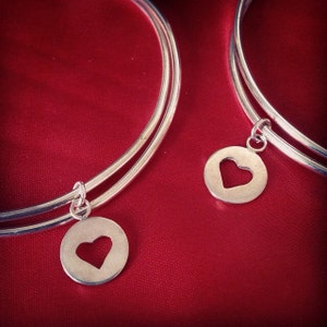 May include: Two silver bangle bracelets with heart-shaped charms. The bracelets are layered on top of each other and are on a red background.