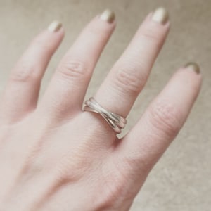 Puede incluir: Un anillo de plata con tres bandas entrelazadas. El anillo está en el dedo de una persona.