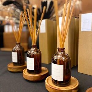 May include: Three brown glass bottles with wooden sticks and labels that read "Ocean Surf & Sea Salt" and "Cedar & Sandalwood" on a wooden surface. The bottles are filled with a clear liquid and are part of a reed diffuser set.