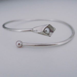 May include: A silver bangle bracelet with a delicate flower design. The flower has a small pearl in the center. The bracelet is open and has a simple, elegant design.