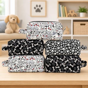 May include: A stack of five rectangular zippered pouches featuring various dog-themed patterns. Two pouches have a white background with black and grey dog illustrations, two have a black background with white bones, and one has a black background with white paw prints.