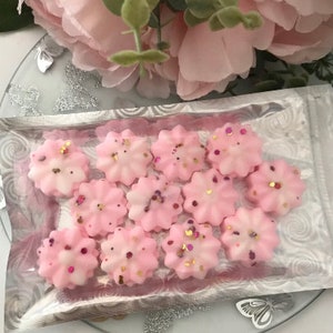 K&ouml;nnte beinhalten: Nahaufnahme einer Packung rosa Wachs-Schmelzen in Form von Blumen. Die Wachs-Schmelzen sind mit Glitzer verziert und in einer durchsichtigen Plastikt&uuml;te verpackt.