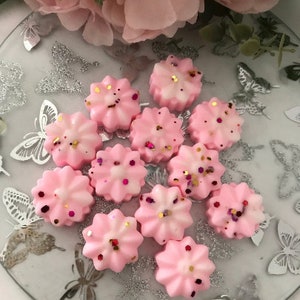K&ouml;nnte beinhalten: Eine Sammlung von rosa Wachs-Schmelzen in Form von Blumen mit goldenen und rosafarbenen Glitzerakzenten. Die Schmelzen sind auf einem silbernen und wei&szlig;en Teller mit Muster angeordnet.