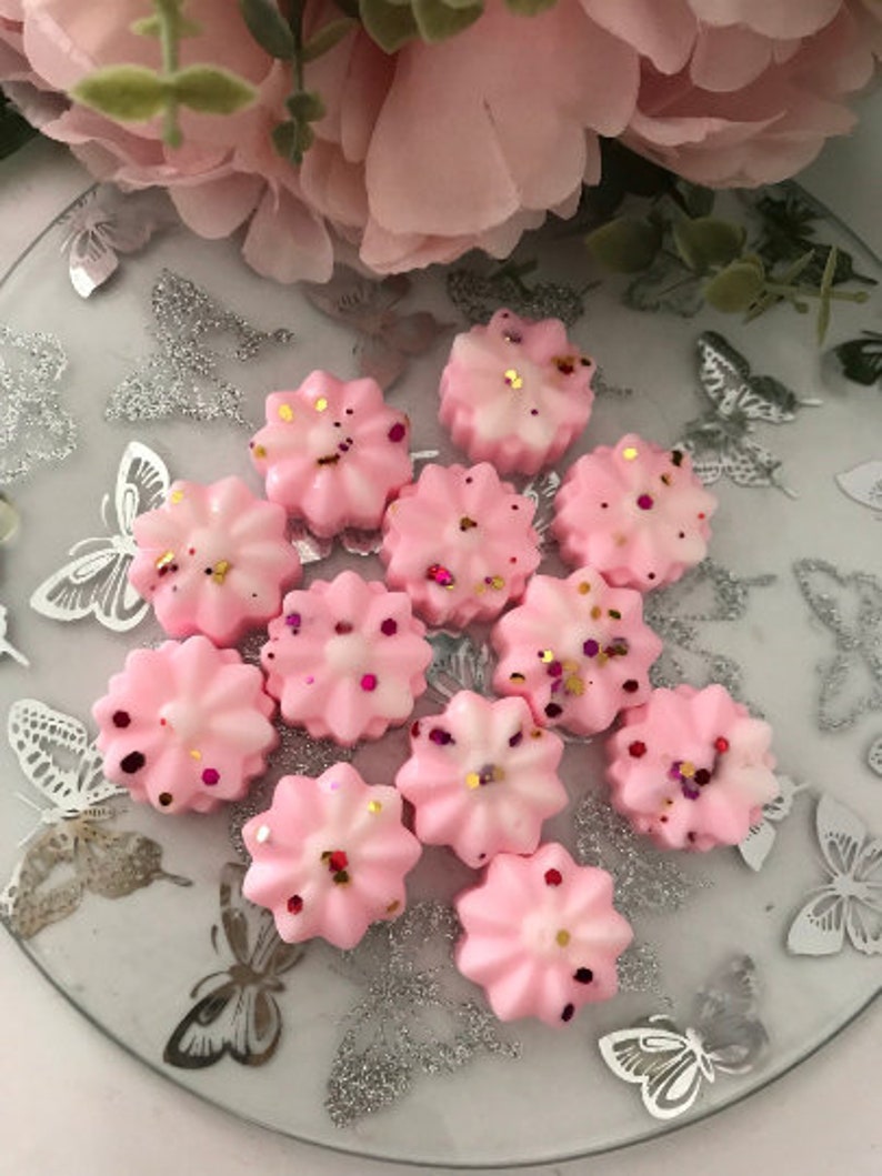 K&ouml;nnte beinhalten: Eine Sammlung von rosa Wachs-Schmelzen in Form von Blumen mit goldenen und rosafarbenen Glitzerakzenten. Die Schmelzen sind auf einem silbernen und wei&szlig;en Teller mit Muster angeordnet.