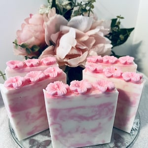 May include: Five bars of handmade soap with a pink and white swirl design, topped with pink flower accents. The soaps are arranged on a clear glass plate with butterfly designs, with a bouquet of pink flowers in the background.