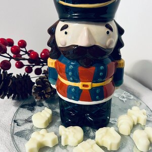May include: A ceramic nutcracker figurine in a soldier's uniform, with a black hat and a brown mustache. The figurine is on a glass plate with white wax melts. Red berries and pine cones are in the background. The nutcracker is a festive holiday decoration.
