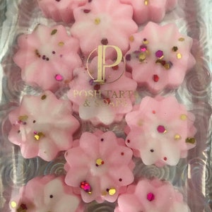 K&ouml;nnte beinhalten: Neun rosa und wei&szlig;e Wachsschmelzen in Bl&uuml;tenform mit goldenem und rotem Glitzer. Die Wachsschmelzen sind in einem durchsichtigen Plastikbeh&auml;lter angeordnet. Der Beh&auml;lter tr&auml;gt den Aufdruck "POSH TARTS & SOAPS".