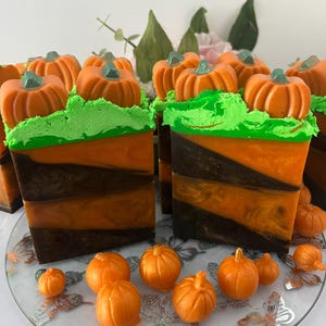 May include: Artisan soap bars with a seasonal autumn theme. The bars have orange and brown layers, topped with green frosting and small orange pumpkin decorations. Additional pumpkin decorations are scattered around the base.