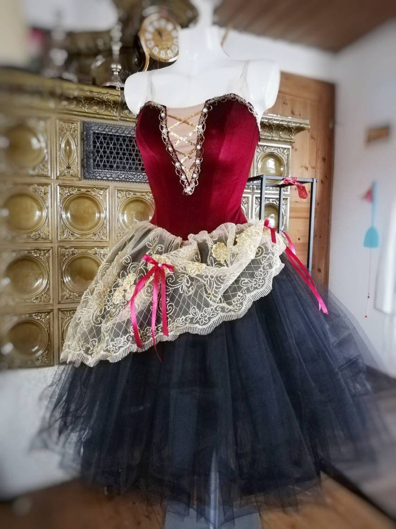 May include: A red velvet bodice with gold and pearl embellishments, a gold and white lace overlay, and a black tulle skirt. The bodice has a sweetheart neckline and a criss-cross design in the front.