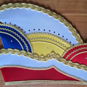 Könnte beinhalten: Ein mehrfarbiger Karnevalshut mit mehrschichtigem Design. Der Hut hat Abschnitte aus weißem, blauem, gelbem und rotem Stoff, die mit goldener Spitze verziert sind. Eine kleine Quaste hängt an der Seite. Strasssteine sind über den Hut verstreut.