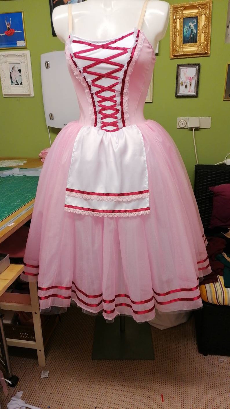May include: A pink tulle ballet tutu with a white satin bodice and red ribbon accents. The bodice has a criss-cross lacing detail and the skirt has a white satin apron with red ribbon trim.
