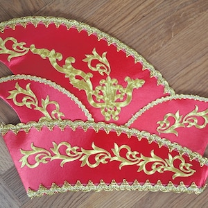 Könnte beinhalten: Roter und goldener Narrenhut mit gewelltem Rand und goldener Verzierung. Der Hut hat goldene, gestickte Blumenmuster. Der Hut ist aus glänzendem rotem Stoff gefertigt und wahrscheinlich für ein Kostüm oder einen festlichen Anlass.