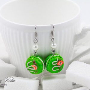 Könnte beinhalten: Handgefertigte grüne Macaron-Ohrringe mit weißen Zuckergussdetails und kleinen roten Blumenakzenten. Jeder Ohrring hängt an einem silberfarbenen Haken mit einer weißen Perle. Die Ohrringe werden in der Nähe einer weißen Tasse und Zuckerwürfeln präsentiert.