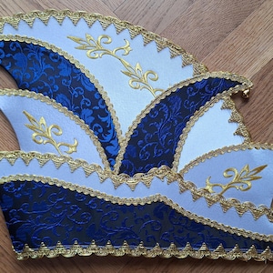 Op de afbeelding: Decoratieve hoed met een gelaagd ontwerp, met witte en marineblauwe panelen, gouden randen en geborduurde accenten. De hoed heeft een geschulpte rand en kleine gouden belletjes aan de uiteinden.