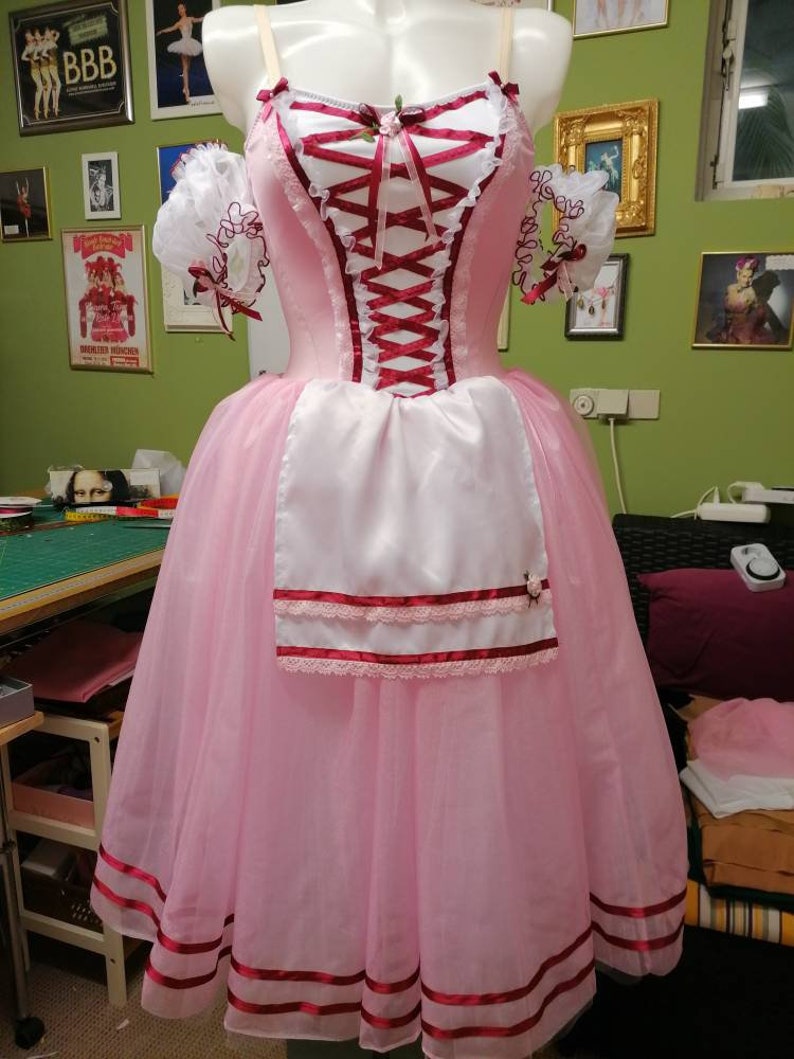 May include: A pink and white ballet tutu with a red ribbon bodice and a white apron. The tutu has a layered skirt with red ribbon trim and a white lace trim.