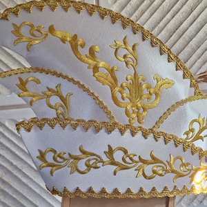Könnte beinhalten: Ein weißer Karnevalshut mit drei markanten Spitzen, verziert mit aufwendigen Goldstickereien und goldener Borte an den Rändern. Kleine goldene Glöckchen sind an den Spitzen befestigt, und lange, dünne, hellbraune Federn ragen an der Rückseite hervor.