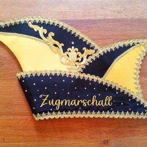 Könnte beinhalten: Eine schwarz-gelbe Narrenkappe mit goldenen Verzierungen und einem goldenen Quasten. Die Kappe hat eine schwarze Satinbasis mit goldenen Pailletten und die Worte "Zugmarschall" in Goldstickerei.