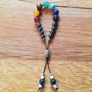 Pode incluir: Um bracelete de contas pretas com um arco-&iacute;ris de pedras coloridas, incluindo verde, azul, amarelo, laranja e vermelho. O bracelete tem uma conta prateada e um cord&atilde;o preto com contas brancas na ponta.