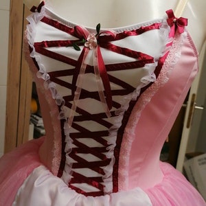 May include: A pink and white ballet tutu with a burgundy bodice featuring a lace trim and a satin ribbon bow with a floral embellishment.