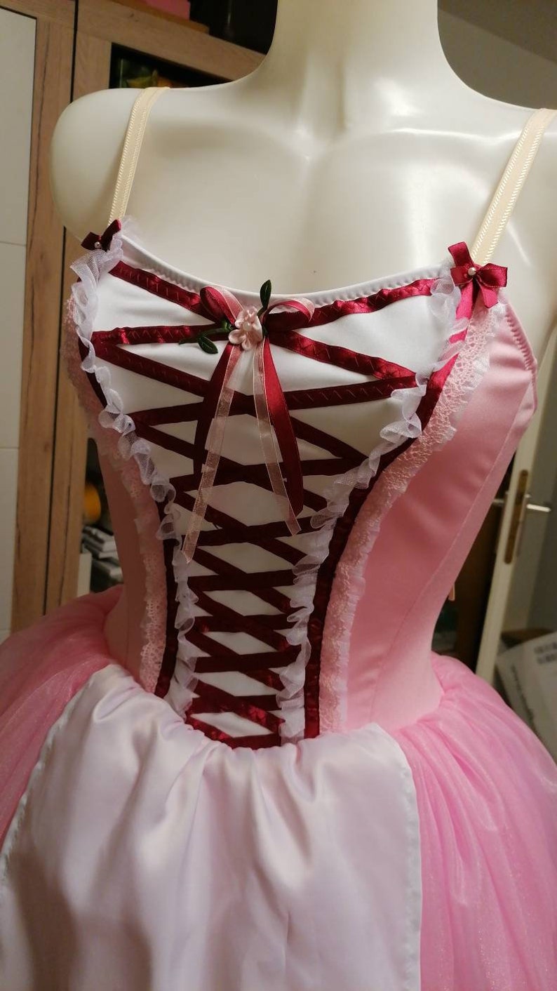 May include: A pink and white ballet tutu with a burgundy bodice featuring a lace trim and a satin ribbon bow with a floral embellishment.