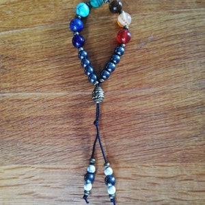 Pode incluir: Um bracelete de contas pretas com um amuleto e uma borla. O bracelete tem sete contas coloridas, incluindo turquesa, azul, verde, laranja, vermelho e marrom.
