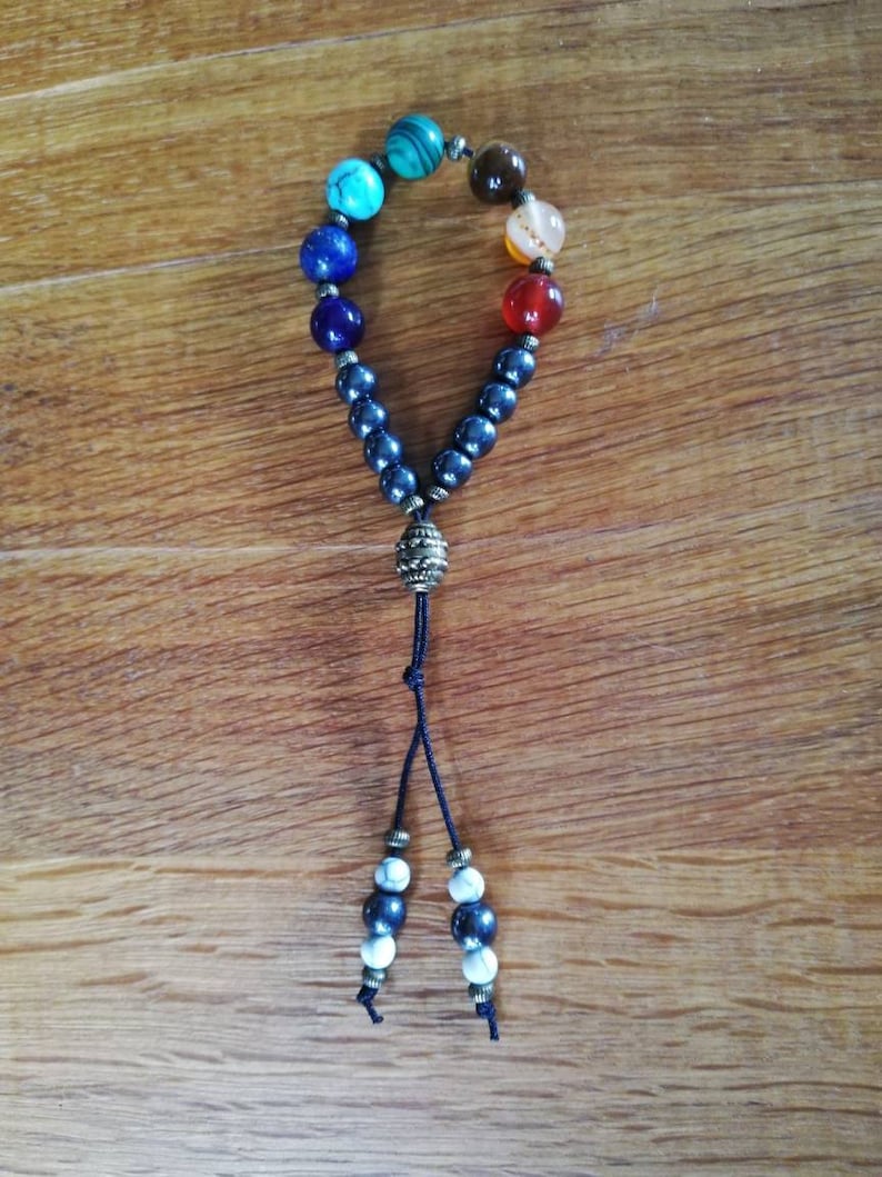 Pode incluir: Um bracelete de contas pretas com um amuleto e uma borla. O bracelete tem sete contas coloridas, incluindo turquesa, azul, verde, laranja, vermelho e marrom.