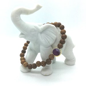 Könnte beinhalten: Ein Armband aus Holzperlen mit einem violetten Edelstein-Akzent. Das Armband wird auf einer weißen Elefantenfigur präsentiert.