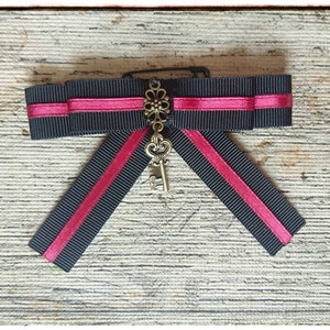 Può includere: Un fiocco di nastro nero e rosa con un ciondolo a forma di chiave decorativo. Il fiocco è legato con un nastro nero e ha una striscia rosa al centro. Il ciondolo a forma di chiave è in metallo e ha un design floreale.