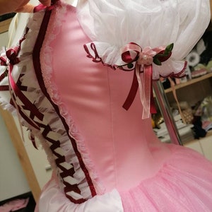May include: A pink and white dress with a white lace bodice and a pink tulle skirt. The dress has a burgundy ribbon and lace trim. The dress has a white lace ruffle on the shoulder.