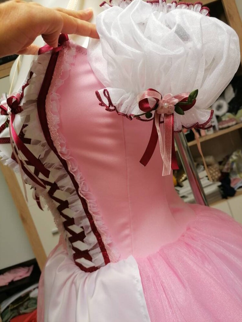 May include: A pink and white dress with a white lace bodice and a pink tulle skirt. The dress has a burgundy ribbon and lace trim. The dress has a white lace ruffle on the shoulder.