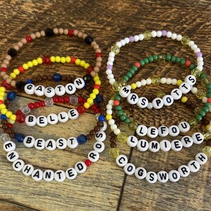 May include: A collection of beaded bracelets with white letter beads spelling out names like "GASTON", "BELLE", and "BEAST". The bracelets feature various colors, including red, yellow, brown, and blue, and are arranged on a wooden surface.