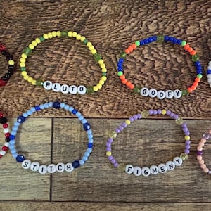 May include: Eight beaded bracelets with character names spelled out in white letter beads. The bracelets are in various colors, including red, yellow, blue, orange, and purple. The names include Mickey, Minnie, Pluto, Goofy, Donald, Stitch, Figment, and Disney.