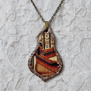 Może przedstawiać: Złoty łańcuszek z zawieszką w postaci błyszczącej, perłowej, brązowo-złotej ramki z obrazem egipskiej kobiety w kolorowej szacie i nakryciu głowy.