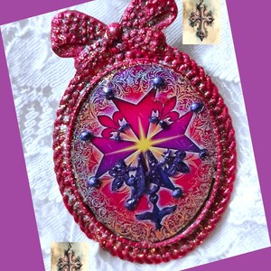 Peut inclure: Une décoration ovale rouge et or avec une finition scintillante. La décoration présente une image colorée d'une croix avec une étoile et une colombe. La décoration est ornée d'un nœud rouge.