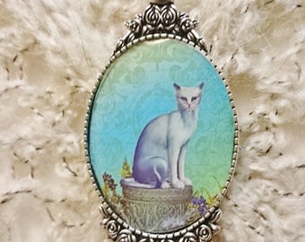 Collier pendentif chat Siamois aux yeux bleu assis sur une margelle de puit, chat blanc sur fond en Camaïeu de vert et de bleu