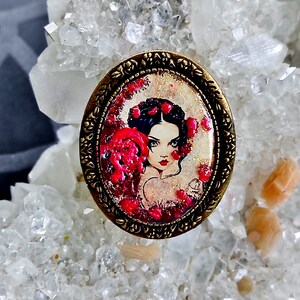 Peut inclure: Une broche ovale de style vintage avec un cadre doré et une image colorée d'une femme aux cheveux noirs et aux fleurs rouges. L'image a une finition scintillante.