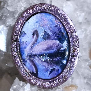 May include: Oval pendant with a miniature painting of a swan on a lake. The artwork is framed in a decorative, ornate, lavender-colored metal. The background is a blue and purple landscape with shimmering details.