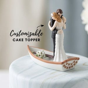 Könnte beinhalten: Eine Tortenaufsatz für Hochzeiten, die ein sich umarmendes Paar in einem kleinen Boot zeigt. Das Boot ist weiß mit braunem Rand und dekorativen Details. Der Text "Customizable CAKE TOPPER" ist ebenfalls sichtbar.