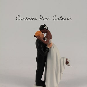 May include: A wedding cake topper featuring a bride and groom embracing. The bride is wearing a white dress and the groom is wearing a black suit. The bride has dark skin and the groom has light skin. The bride is holding a white flower in her hair.