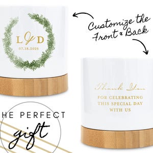 Op de afbeelding: Wit keramisch cadeau met een bamboe basis. De voorkant heeft een groene krans met gouden initialen en de datum 07.18.2026. De achterkant zegt "Thank You for Celebrating This Special Day With Us". De tekst "Customize the Front & Back" is ook zichtbaar.