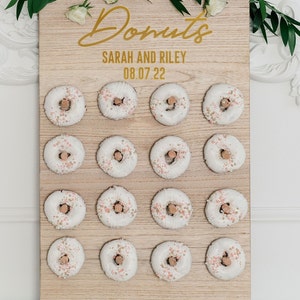May include: A wooden donut wall with the text "Donuts" in gold script, "Sarah and Riley" and "08.07.22" in gold lettering. The wall is decorated with 18 white donuts with pink and gold sprinkles.
