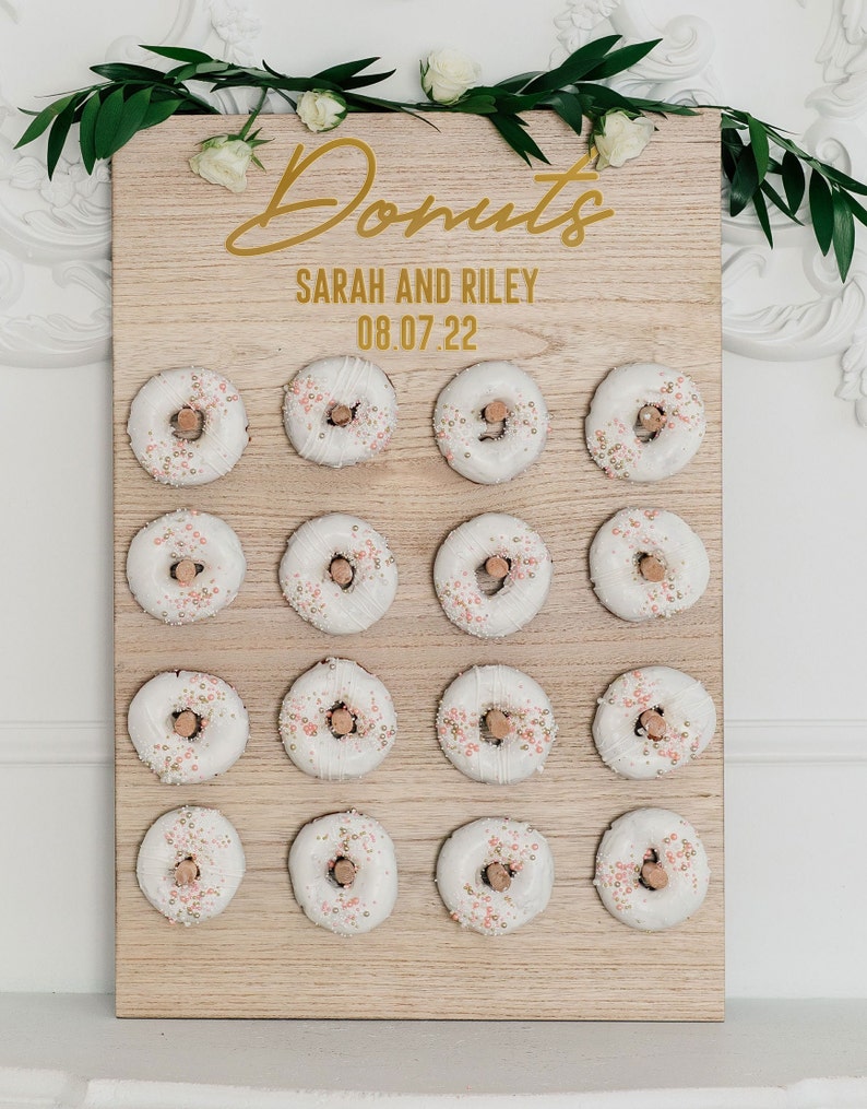 May include: A wooden donut wall with the text "Donuts" in gold script, "Sarah and Riley" and "08.07.22" in gold lettering. The wall is decorated with 18 white donuts with pink and gold sprinkles.